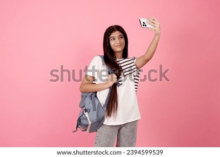 Young smiling teenage girl taking a selfie with phone with bag on pink background in studio.