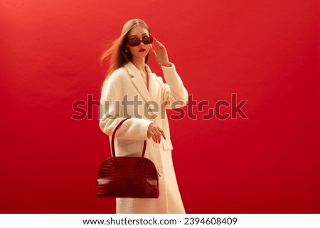 Fashionable confident woman wearing trendy white boucle midi coat, stylish sunglasses, holding faux crocodile leather bag, handbag, posing on red background. Copy, empty space for text Royalty-Free Stock Photo #2394608409