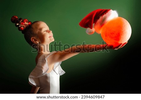A cute little girl gymnast in a white leotard turned sideways to the camera, holding a red glowing ball in her outstretched hands. The ball is wearing a Santa Claus hat. Green background.