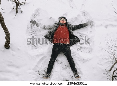 A stylish, fashionable man in sunglasses lies, rests on the white snow in winter and moves his arms like a bird in a snowfall. Photography, concept. Shooting a motion picture.
