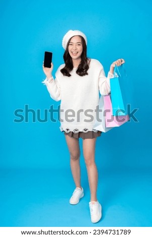 Full body photo of young Asian woman with long hair, online business shopper Carrying several shopping bags, smiling holding a smartphone wearing a white coat Photographing a blue screen in a studio