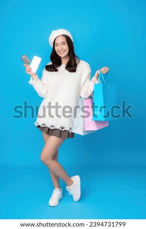 Full body photo of young Asian woman with long hair, online business shopper Carrying a credit card and a shopping bag, a smartphone, wearing a white coat. Photographing a blue screen in a studio