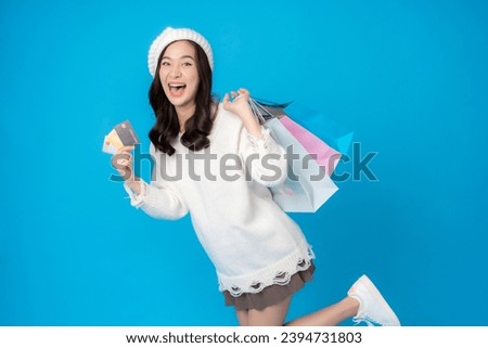 Asian female online shopper with long hair happy and laughing Holding a credit card and a shopping bag, wearing a coat and hat, taking photos against a blue backdrop in the studio.