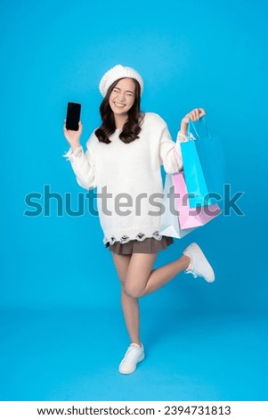 Full body photo of young Asian woman, online business shopper, excited, holding a smartphone, long hair, wearing a white coat. Carrying several shopping bags Photographing a blue screen in a studio