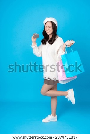 Full body image of young Asian woman with long hair, online business shopper. Holding a credit card and a smartphone There are shopping bags. Wearing a coat to photograph a blue backdrop in a studio