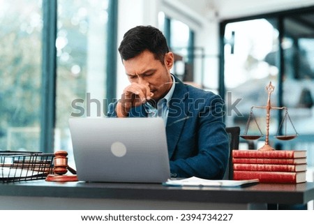 Asian male business lawyer looks stressed, pinching the bridge of his nose in front of a laptop at his office desk.
