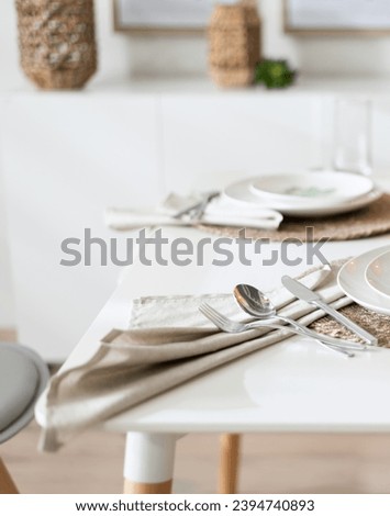 Luminous Minimalist Dining with Sleek White Tableware on Woven Placemats, Soft Linen Napkins, Polished Silverware, All on a Modern Pristine White Dining Table Bathed in Natural Light, Wooden Flooring. Royalty-Free Stock Photo #2394740893