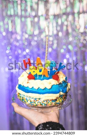 A hand holds up a colorful birthday cake against a unicorn-colored background.