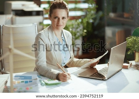 Portrait of modern middle aged woman worker in a light business suit in modern green office with documents and laptop.