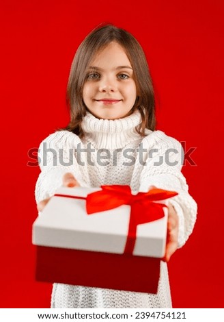 Child with arms extended forward holding gift box with red ribbon and bow. Little girl looking at camera. Kid wearing white sweater on red background, focus on foreground