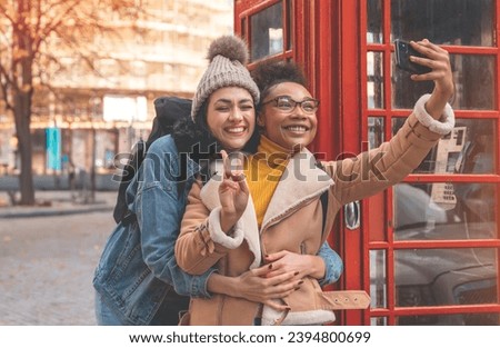 two happy friends, girlfriend and women using a mobile phone, camera and taking a selfie against a red phonebox in a city in England. Travel Lifestyle concept