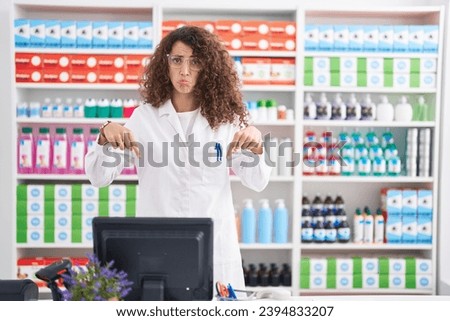 Hispanic woman with curly hair working at pharmacy drugstore pointing down looking sad and upset, indicating direction with fingers, unhappy and depressed.  Royalty-Free Stock Photo #2394833207