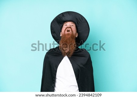 Young wizard in halloween isolated on blue background looking up and with surprised expression