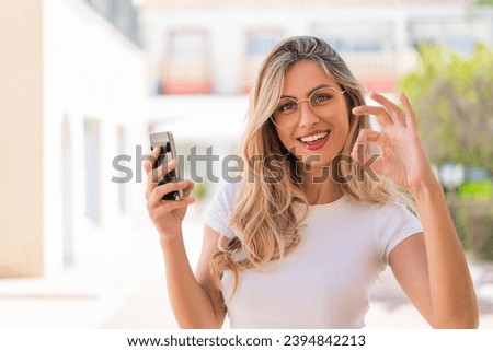 Pretty blonde Uruguayan woman using mobile phone and doing OK sign