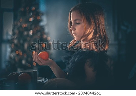 a cute girl in a black dress sits at a table with tangerine on the background of the Christmas tree. holiday, new year, christmas