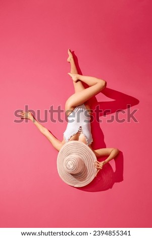 Stylish, elegant young woman in swimsuit and straw hat lying against pink studio background. Sunbathing, resting. Concept of summer, vacation, resting, holidays, fashion and beauty