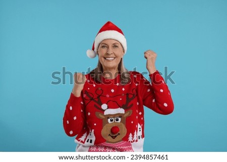 Happy senior woman in Christmas sweater and Santa hat on light blue background Royalty-Free Stock Photo #2394857461