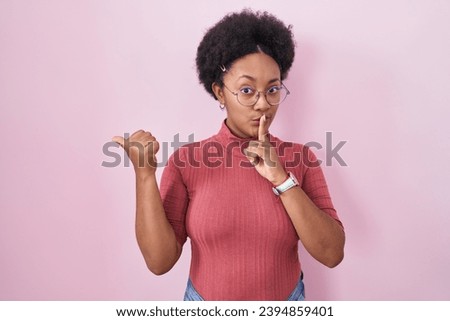 Beautiful african woman with curly hair standing over pink background asking to be quiet with finger on lips pointing with hand to the side. silence and secret concept. 