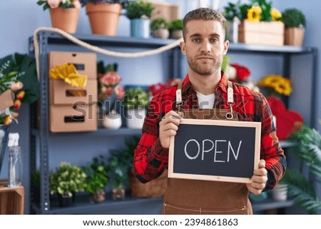 Young caucasian man working at florist holding open sign relaxed with serious expression on face. simple and natural looking at the camera. 