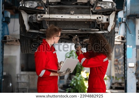 car lift under vehicle. Caucasian manager use laptop computer and hispanic latin male mechanic repairs car in garage. Car repair maintenance and auto service garage concept