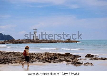 Tourists wearing wide-brimmed straw hats and no shoes walk in the beautiful sea and look at a distant lighthouse. A woman carrying a red backpack, yellow shirt and hat looks at lighthouse in Khao Lak