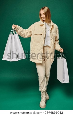 full length of cheerful blonde woman in trendy winter attire holding white shopping bags on green