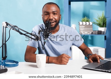Smiling vlogger speaking in microphone, using laptop and looking at camera while recording video for internet channel. Influencer blogger talking in mic while streaming online portrait