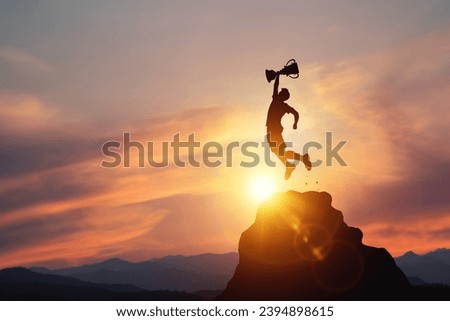 Silhouette of a businessman jumps holding a trophy on top mountain with light sunset. concept of a successful business or determination to lead the organization to success. Royalty-Free Stock Photo #2394898615
