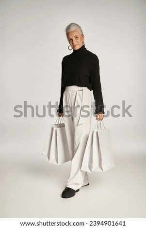 happy middle aged woman in elegant attire and hoop earrings standing with shopping bags on grey