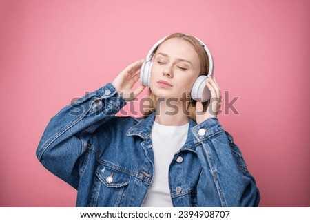 Happy girl listening to music with wireless big headphones on pink background.
