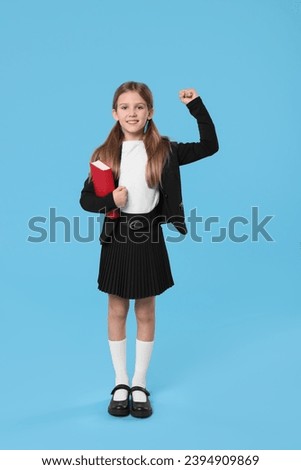 Happy schoolgirl with book on light blue background Royalty-Free Stock Photo #2394909869