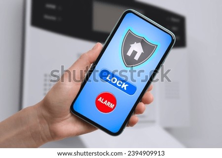 Woman operating home alarm system via mobile phone against white wall with security control panel, closeup. Application interface on device screen