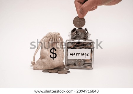 A person putting a coin into a glass jar  with word "marriage" on paper post it. A small pouch bag sitting next to glass jar and a pile of coins. Saving and finance concept