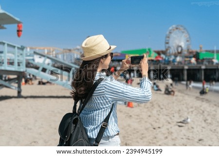 rear view of asian Japanese girl backpacker taking photos with smartphone on beach of santa monica pier near Lifeguard Tower