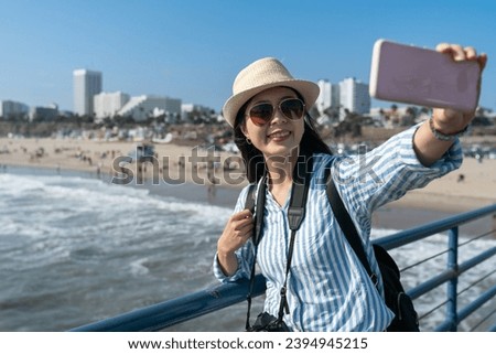 cheerful asian korean girl visitor holding phone and taking selfie picture at railing with popular beach at background. travel lifestyle and people on holiday