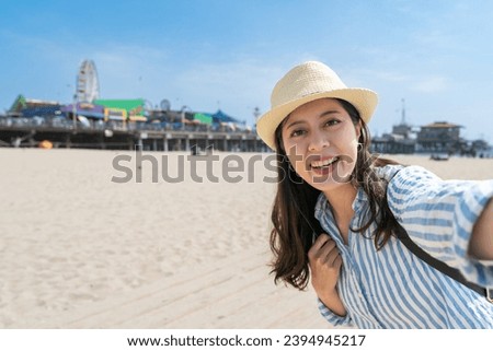 cheerful asian Taiwanese woman visitor smiling at camera while taking selfie picture on santa monica sand beach with Ferris Wheel at back on sunny day