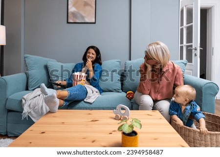 Young irresponsible mother watch TV and eat popcorn while her mother child grandmother looks after the baby. Carefree mom pays no attention to her child who is being looked after by older babysitter Royalty-Free Stock Photo #2394958487