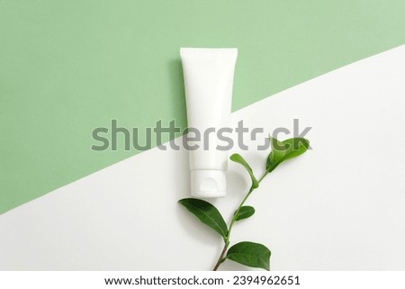 An unlabeled plastic tube is displayed with fresh green tea leaves on a pastel two-color background. Tannins in green tea act as an astringent to shrink pores. Royalty-Free Stock Photo #2394962651