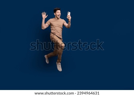 Full size photo of eccentric man wear beige outfit jumping talk on smartphone video call say hi isolated on dark blue color background
