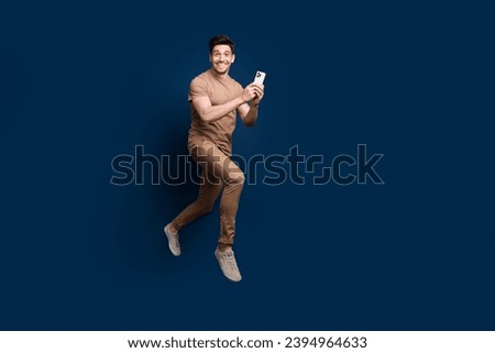 Full size photo of positive man wear beige outfit jumping hold smartphone enjoy fast speed internet isolated on dark blue color background