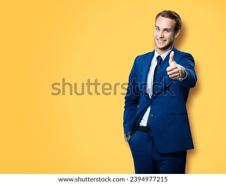 Excited businessman showing thumb up like sign gesture, in blue confident style suit, against vivid yellow background. Handsome happy man. Copy space for slogan or text.