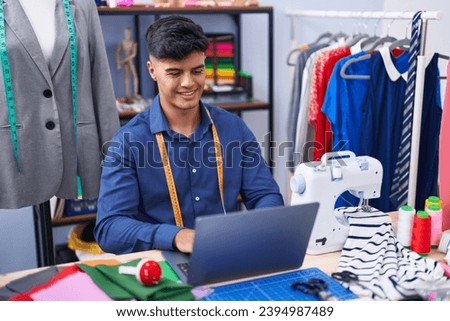 Young hispanic man tailor smiling confident using laptop at clothing factory