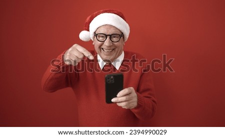 senior wearing christmas hat pointing to smartphone over isolated red background