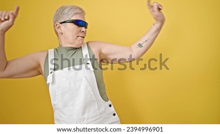 Middle age grey-haired woman using virtual reality glasses dancing over isolated yellow background