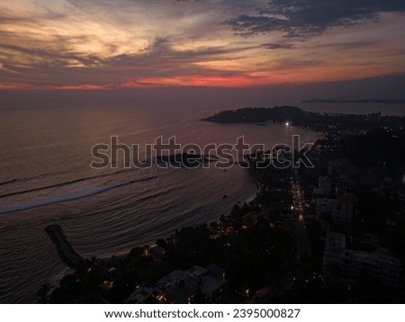 Drone capture of coastal cityscape at dusk with vibrant sunset sky. Urban shoreline illuminated as day transitions to night. Ocean view on sunset. Sunrise above sea. Mirissa, Sri Lanka. Aerial.