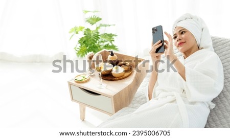 Relax young Asian woman in bathrobe, towel on a head, lying on the sofa bed while using smartphone and beauty treatment items for spa procedures such as essential oil and salt massage.