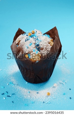 Cupcake with icing sugar and sprinkles on a blue background, vertical photo, close-up. Backdrop with a decorated cupcake wrapped in brown butter paper, vertical picture.