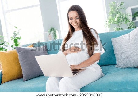 Photo of cute lovely adorable pregnant girl future mommy sitting on couch touching tummy choosing clothes for baby flat indoors