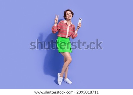 Full size photo of cheerful nice positive woman dressed print shirt holding smartphone showing v-sign isolated on blue color background
