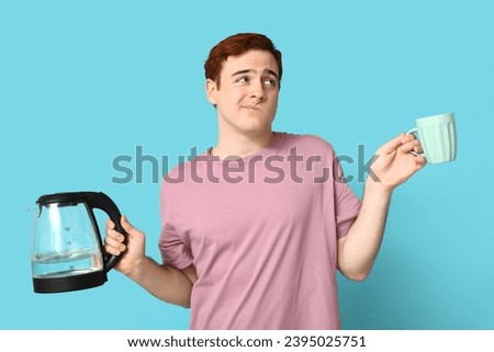Confused young man with modern electric kettle and cup on blue background